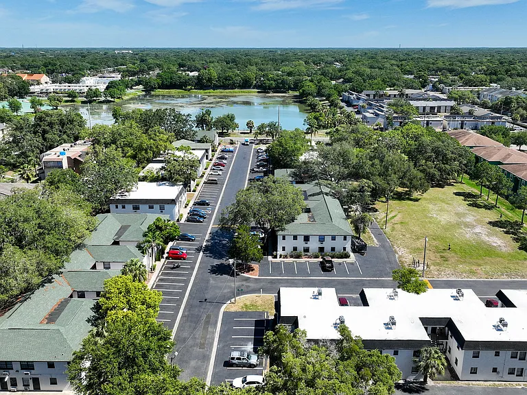 Scenic aerial view of a charming apartment complex surrounded by lush greenery and serene water views, showcasing spacious parking areas and well-maintained buildings.