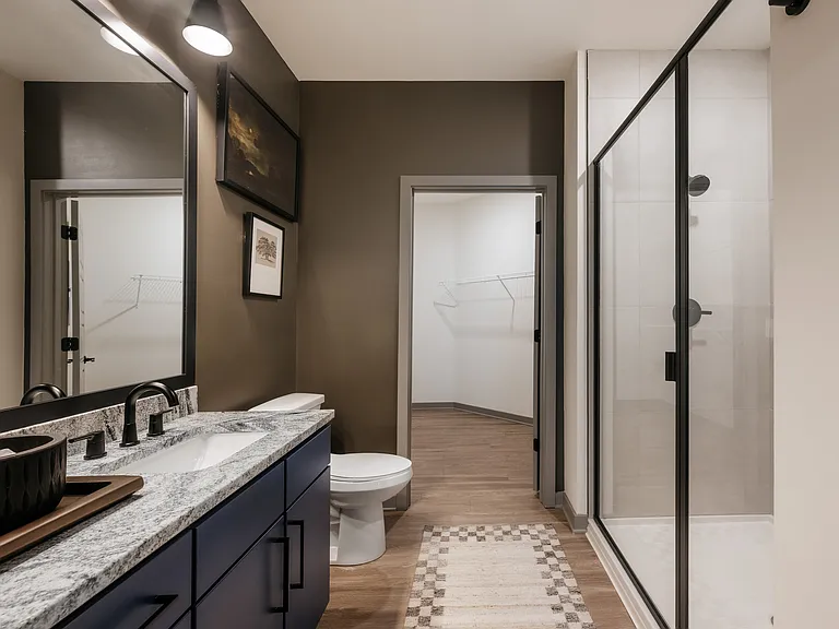 Contemporary apartment bathroom with wood-inspired plank flooring, granite countertops, and semi-frameless glass shower with tile surround.