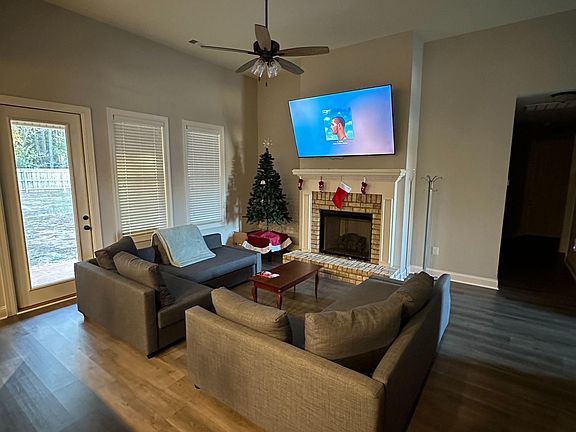 Spacious living room with a fireplace, ceiling fan, and a seating area centered around a wall-mounted TV and large windows.