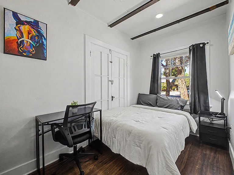 Private bedroom with a full bed, workstation desk, and nightstand, plus a window with curtain panels for natural light.