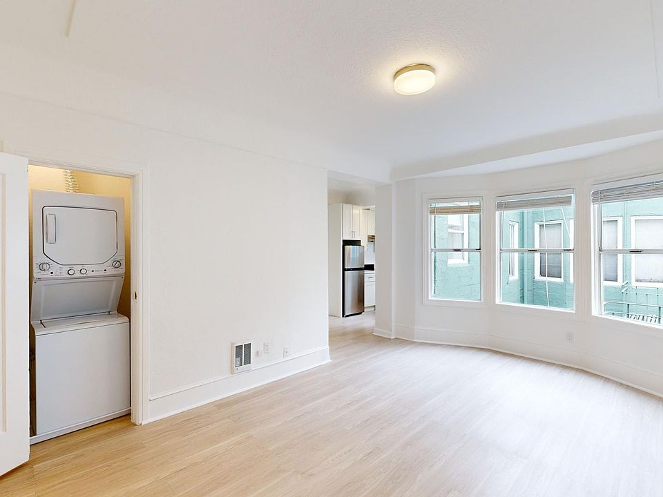 Bright, airy room featuring a stackable washer-dryer, large windows, and wood flooring, leading into a kitchen area.