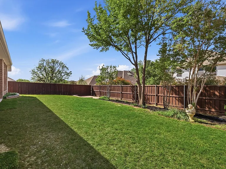 Spacious grassy backyard featuring a full privacy fence, mature trees, and a brick exterior building wall
