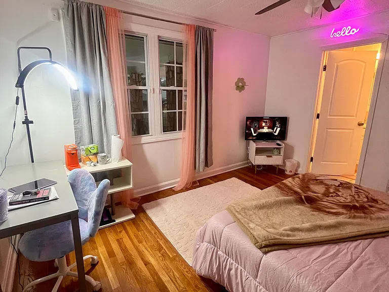 Furnished bedroom with hardwood floors, ceiling fan, and a work desk, plus a window with curtains and a TV stand.