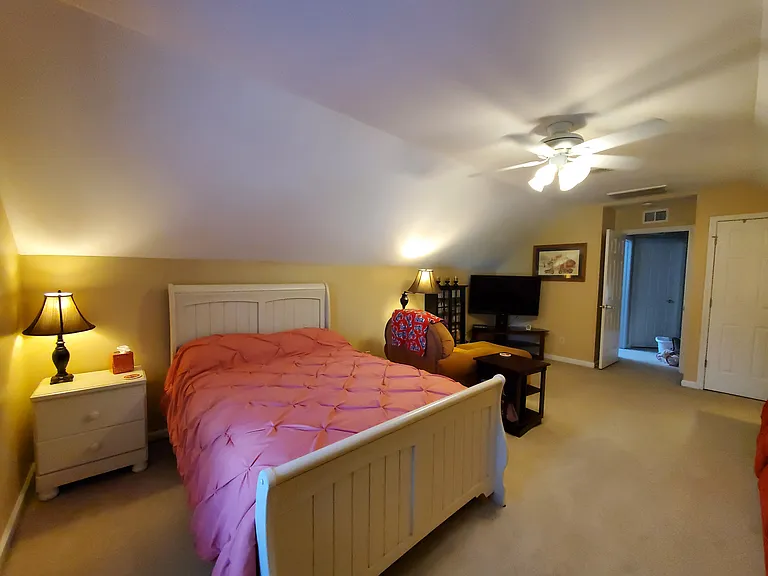 Full size bed with mattress and box springs, nightstand, lamp. Behind that is an overstuffed chair and ottoman and corner TV stand.