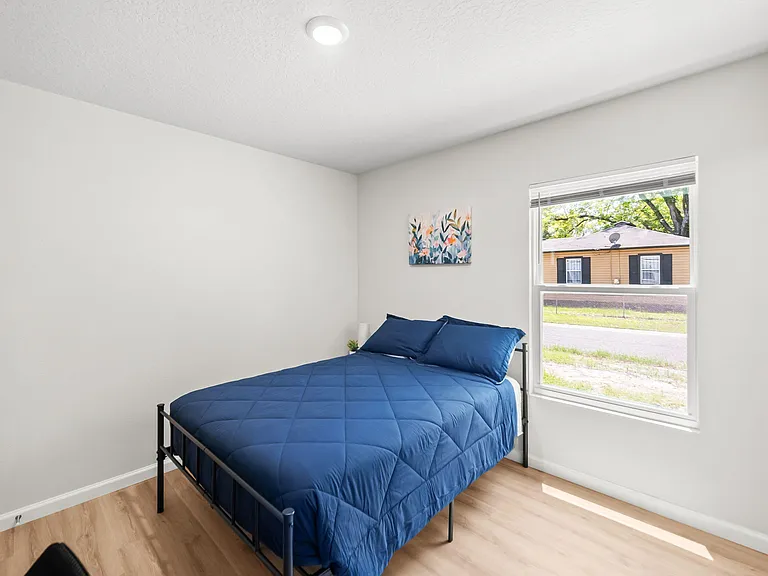 Private bedroom with a full-size bed, wood-look flooring, and a large window with blinds for natural light and ventilation.