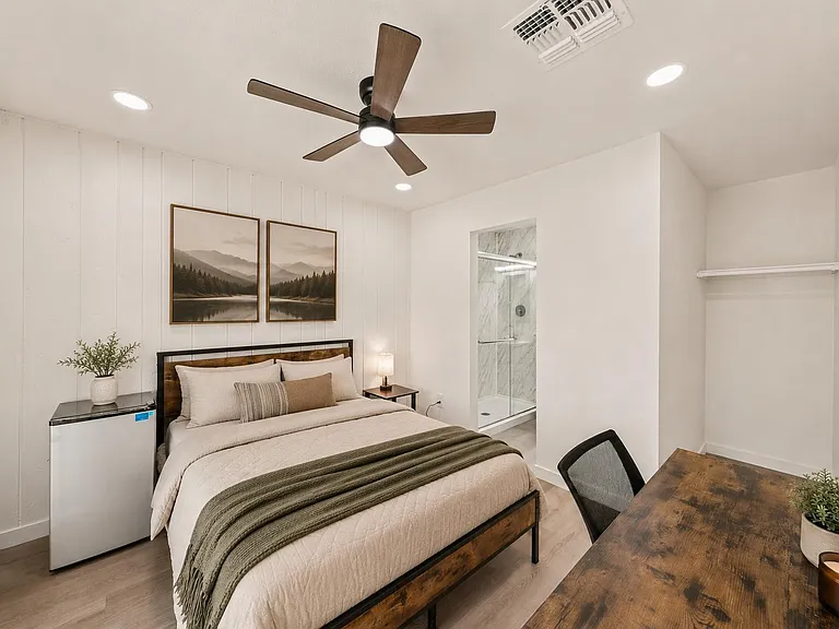 Furnished bedroom with ceiling fan, mini fridge, work desk, and an en-suite bath with a glass shower enclosure.