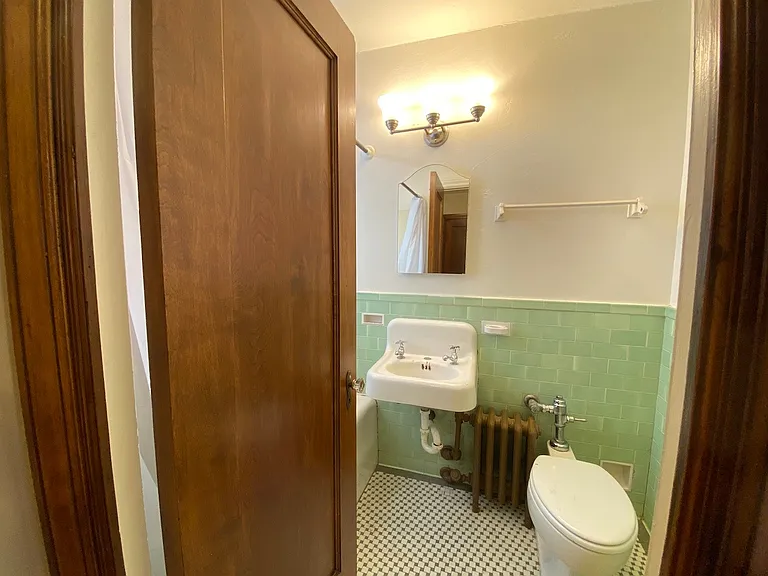 The bathroom blends preserved historic charm with crisp vintage styling, featuring original mint green tilework, mosaic floors, warm wood trim, and classic fixtures that give the space a timeless and refreshing feel.
