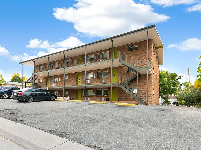 Charming brick apartment building with vibrant green doors, surrounded by lush trees and ample parking, under a clear blue sky.