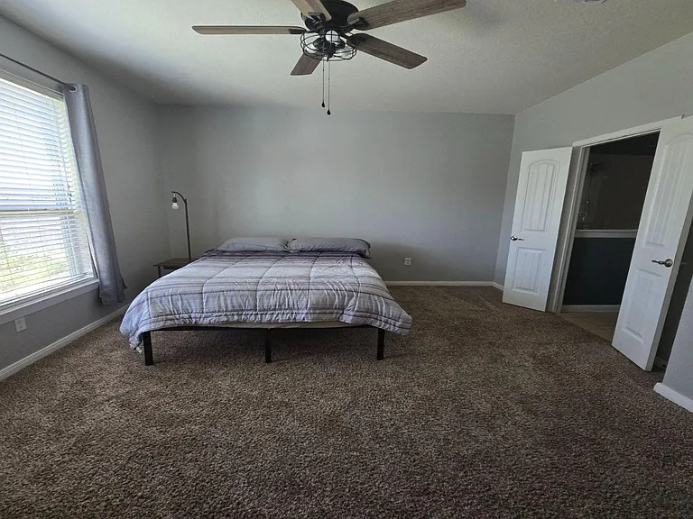 Spacious carpeted bedroom with a large window, ceiling fan, and convenient closet access for everyday storage.