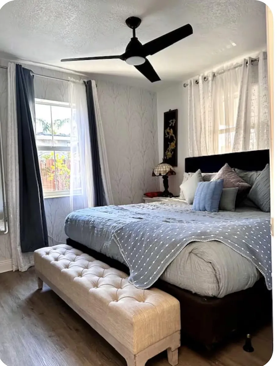 Bedroom with a queen bed, ceiling fan, bedside table, and two windows with curtain panels for natural light. - SW 2nd Ave
