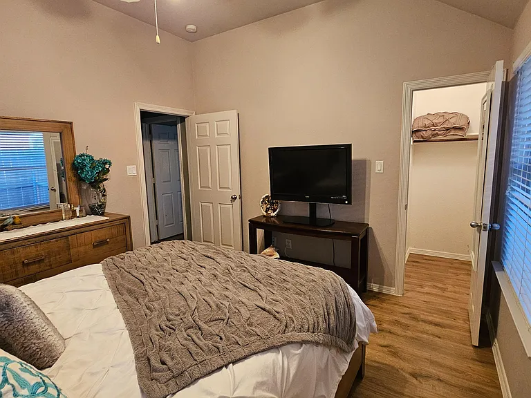 Furnished bedroom with a bed, dresser, and TV, plus a large window with blinds and easy access to closet storage.