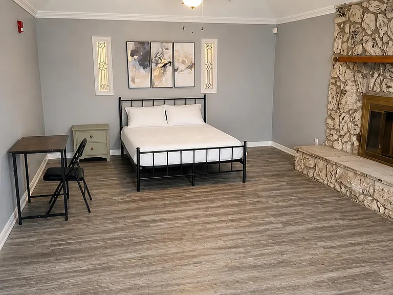 Spacious furnished bedroom with wood-look flooring, ceiling fan, workspace, and a decorative fireplace feature wall.