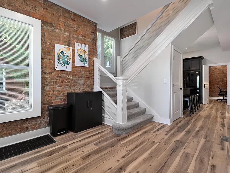 Welcoming entry hallway with exposed brick accent wall, durable wood-look flooring, and a carpeted staircase leading upstairs