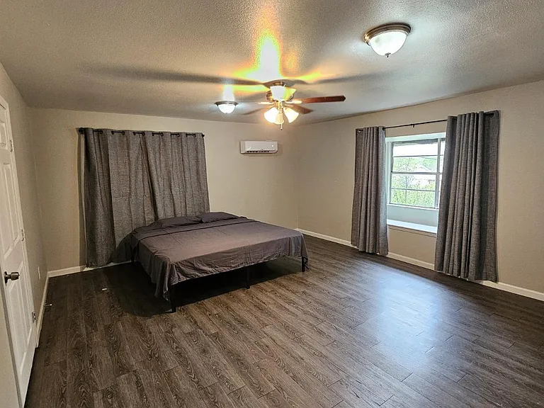 Spacious bedroom with wood-look floors, ceiling fan, mini-split A/C, and a large window with full drapery panels.