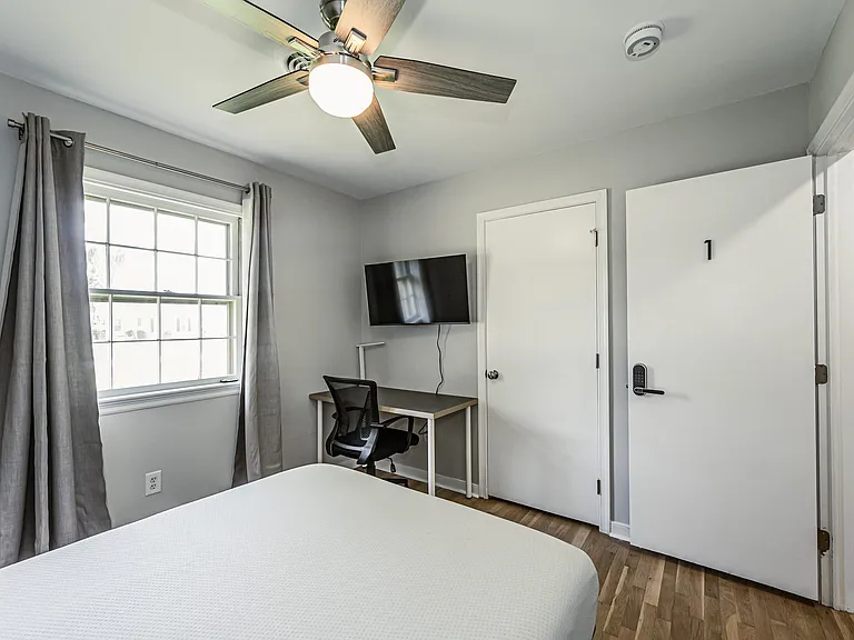 Bedroom with a large window, ceiling fan, wall-mounted TV, and a compact desk area for working or studying.