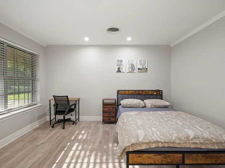 Spacious furnished bedroom with a large window, desk workspace, and bedside storage for everyday convenience.