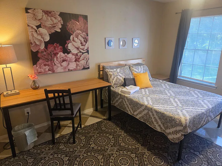 Furnished bedroom with a full-size bed, desk workspace, and a large window with blinds for natural light and privacy.
