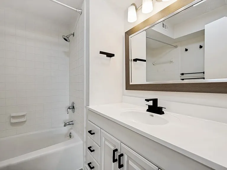 Bathroom featuring a white bathtub with subway tile surround, white vanity with integrated sink, black modern faucet, and a framed mirror with overhead lighting