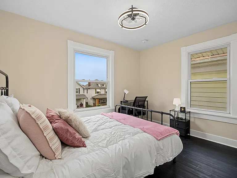 Furnished bedroom with a full bed, two windows for natural light, and a dedicated desk setup for work or study.