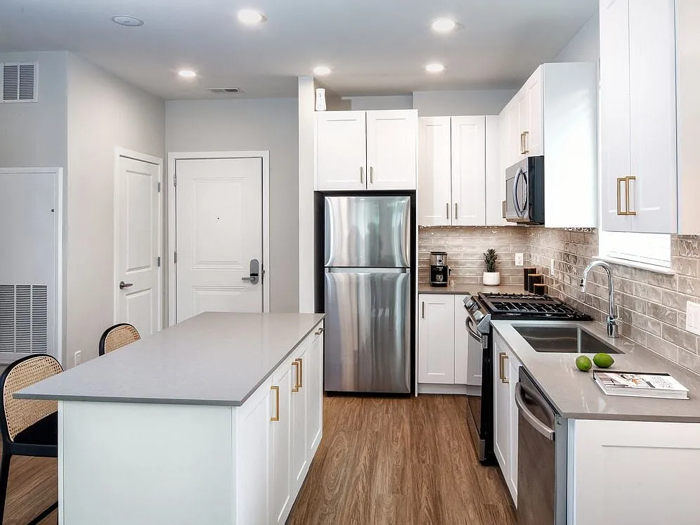 Design Collection II (South Building) Kitchen with white cabinetry, gold pulls, beige tile backsplash, grey quartz countertops, stainless steel appliances, and hard surface flooring - Avalon Somerville Station