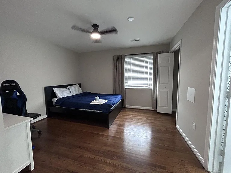 Spacious bedroom with hardwood floors, ceiling fan, and a large window with blinds; includes closet storage and room for a de