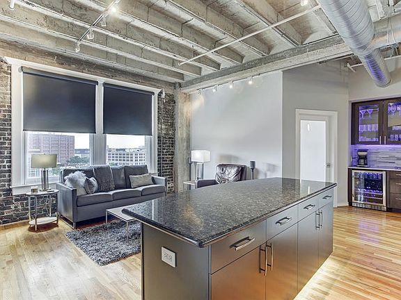A view of the living room from the kitchen. The door to the left of the dry bar leads to the secondary bedroom. Motorized window shades on all windows and notice the historic over 100 year old brick walls.