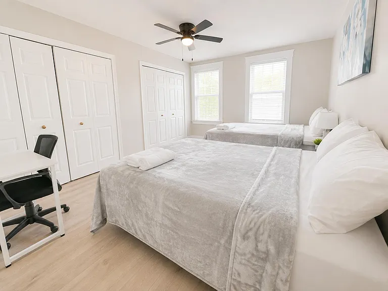 Spacious shared bedroom with two beds, large closets, ceiling fan, and a workstation area beside sunny windows.