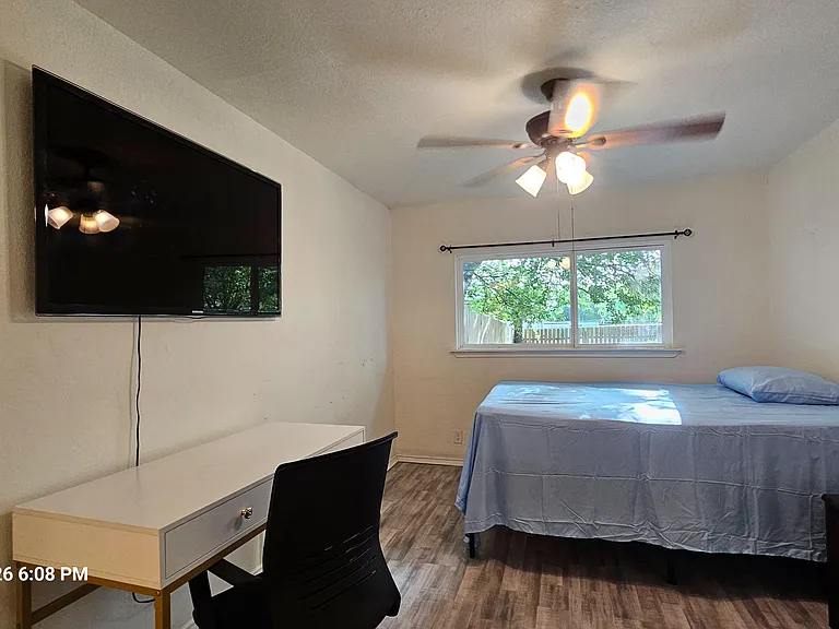 Private bedroom featuring a bed, dedicated desk space, wall-mounted TV, ceiling fan, and a large window overlooking the yard.