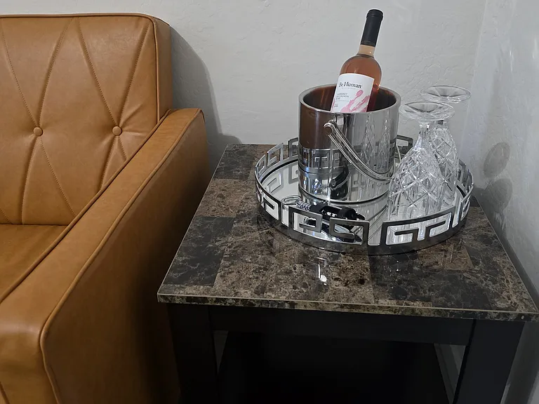 A cozy lounge corner features a marble-top side table with a silver tray offering a bottle of wine, crystal glasses, and a wine opener. Paired with a cushioned leather chair, this elegant touch invites guests to unwind in comfort and style.