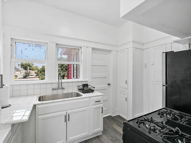Functional kitchen with tiled counters, large sink, gas range, refrigerator, and cabinetry plus window-side prep space.