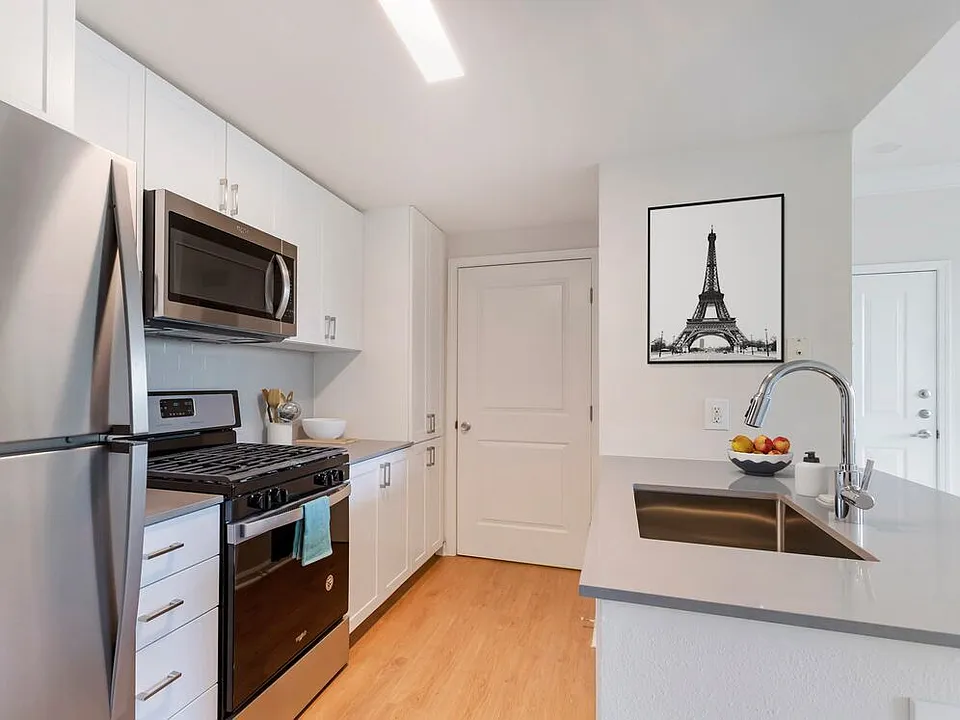 Finish Package II kitchen with white cabinetry, grey quartz countertops, extended hard surface flooring in select units, stainless steel appliances, and tile backsplash