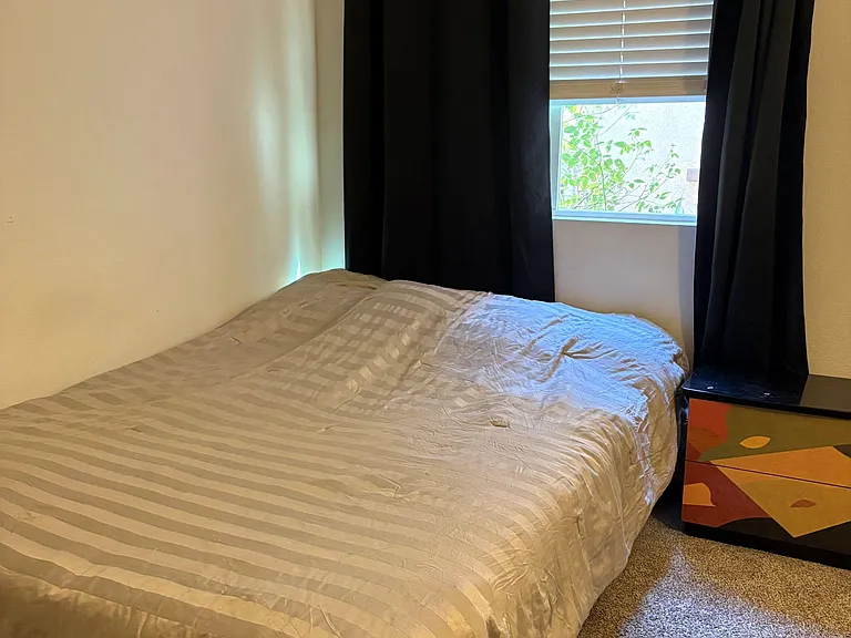 Private bedroom featuring a full-size bed, carpeted flooring, and a window with blinds and blackout curtains for light contro