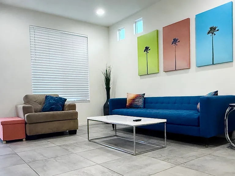 The fun fully-furnished living room has a ceiling fan, sofa, chairs, end table, 65in TV, access to balcony, and view of the kitchen/dining!
