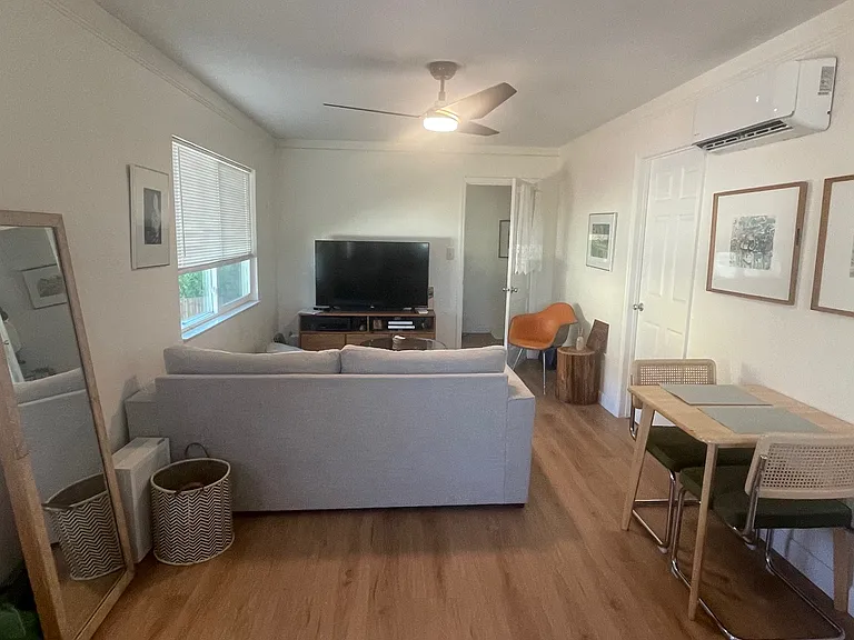 Living room, taken from the kitchen area, shows mini-split A/C unit, ceiling fan, and large window with blinds.