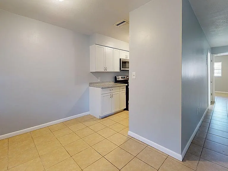 Bright kitchen area with sleek white cabinets, modern appliances, and inviting tiled flooring, leading towards sunlit corridors.