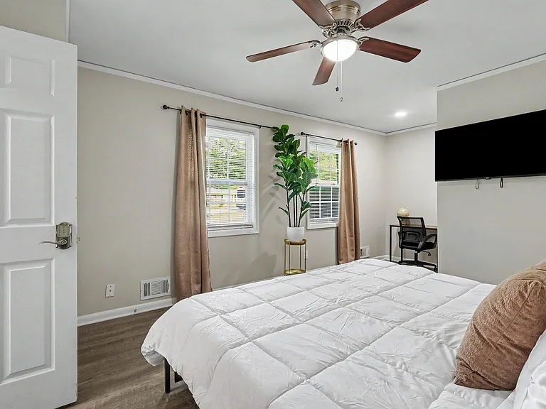 Furnished bedroom with a ceiling fan, two windows with curtains, and a wall-mounted TV plus a compact work desk.