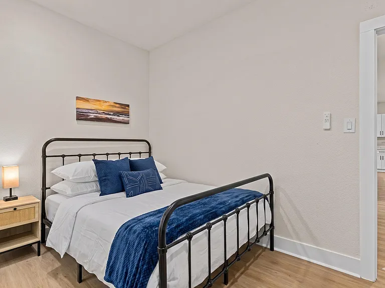 Furnished bedroom with a full bed, nightstand, and lamp, plus easy access to the nearby hallway and kitchen.