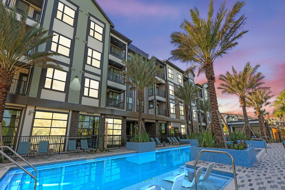 Modern apartment building with stylish balconies overlooking a luxurious pool and palm trees under a vibrant sunset sky.