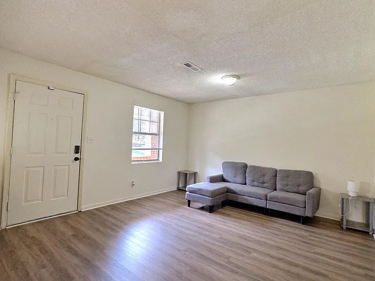 Spacious living room with wood-look flooring, a comfortable seating area, and a window for natural light near the entry.