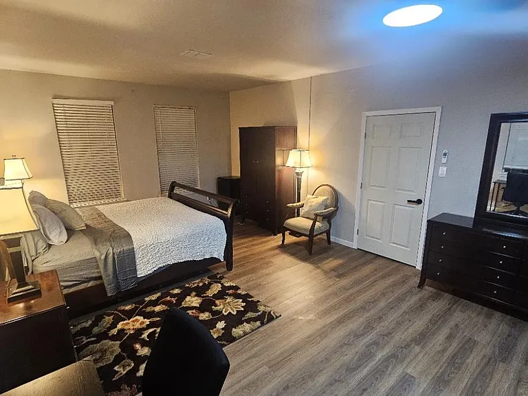 Furnished bedroom with queen bed, nightstands, dresser, and lounge chair, plus window blinds and wood-look flooring.