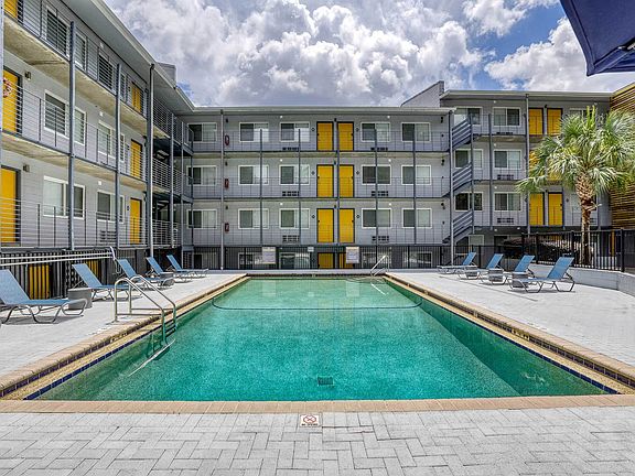Modern apartment complex with a pristine swimming pool, surrounded by stylish lounge chairs and vibrant yellow accents, under a bright, cloudy sky.