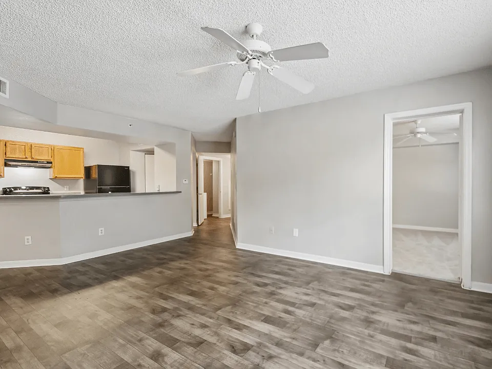Open living room with wood-style flooring, ceiling fan, kitchen breakfast bar, and bedroom access in a 3-bedroom, 2-bath apartment at Gregory Cove in Jacksonville, FL.