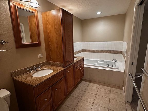 Spacious primary bathroom featuring dual vanities with granite countertops, abundant cabinet storage, and tile flooring throughout. Includes a whirlpool tub, separate walk-in shower, and bright overhead lighting for a clean, functional layout.