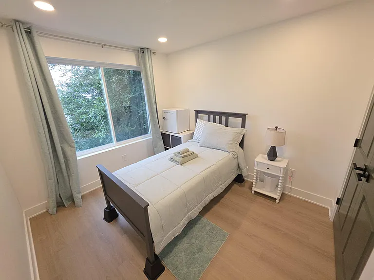Private furnished bedroom with a large window, bedside table and lamp, plus a mini fridge for added convenience.