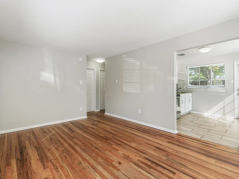 Bright and inviting living area with elegant hardwood floors leading into a sunlit kitchen featuring modern cabinetry and large windows.