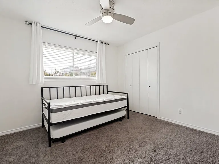 Bright bedroom featuring a large window with blinds, a ceiling fan with integrated lighting, soft gray carpeting, and a double-door closet