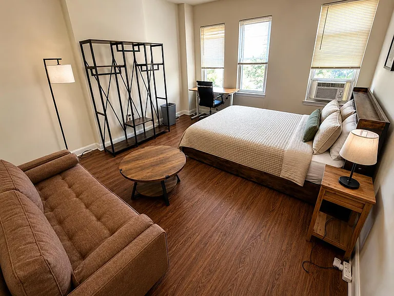 Spacious furnished bedroom with a work desk, open shelving, and large windows with in-room A/C.