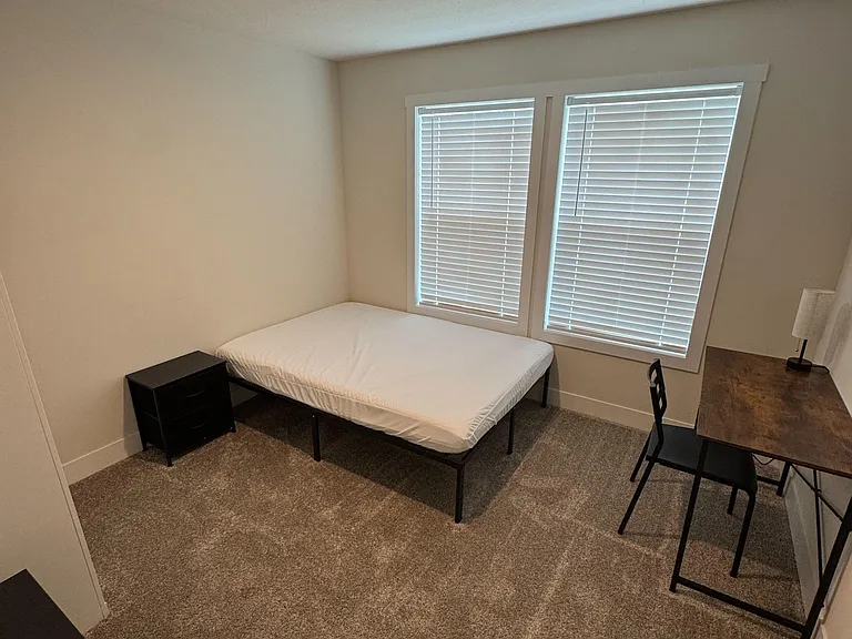 Furnished bedroom with a full-size bed, desk workspace, and large window with blinds for light and privacy.