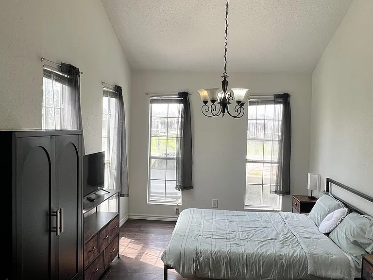 Spacious bedroom with dark wood floors, three large windows, a bed, TV setup, and ample storage with dresser and armoire.