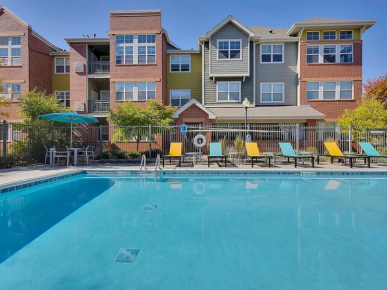 Modern apartment complex with a vibrant pool area, featuring colorful lounge chairs and shaded seating, perfect for relaxation and leisure.
